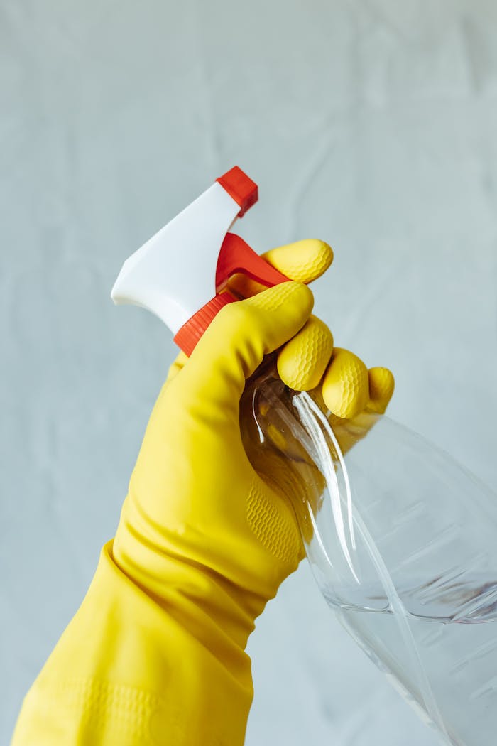 services-03 A hand wearing a yellow rubber glove holds a red and white spray bottle against a neutral background.