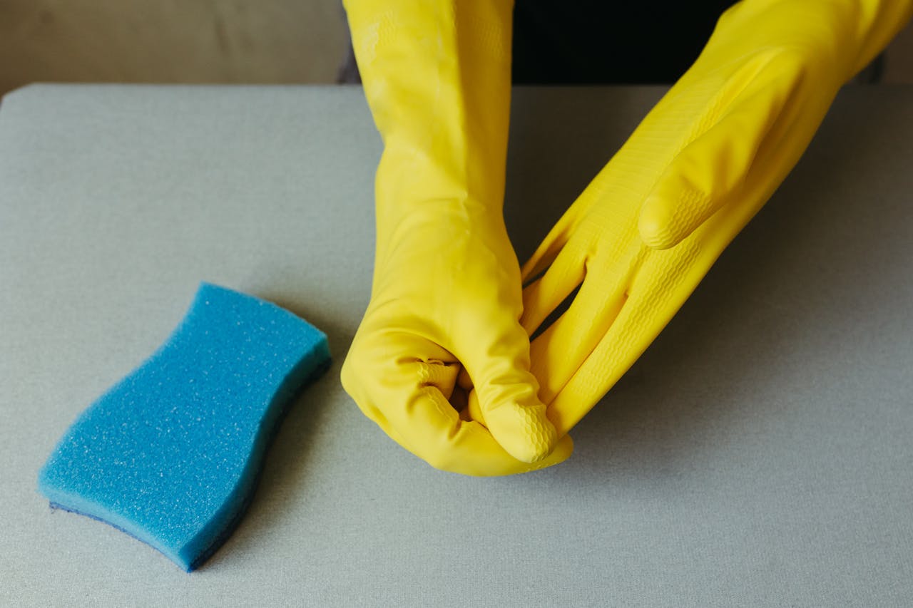 gallery-6 Close-up of hands wearing yellow gloves next to blue sponge on gray surface.