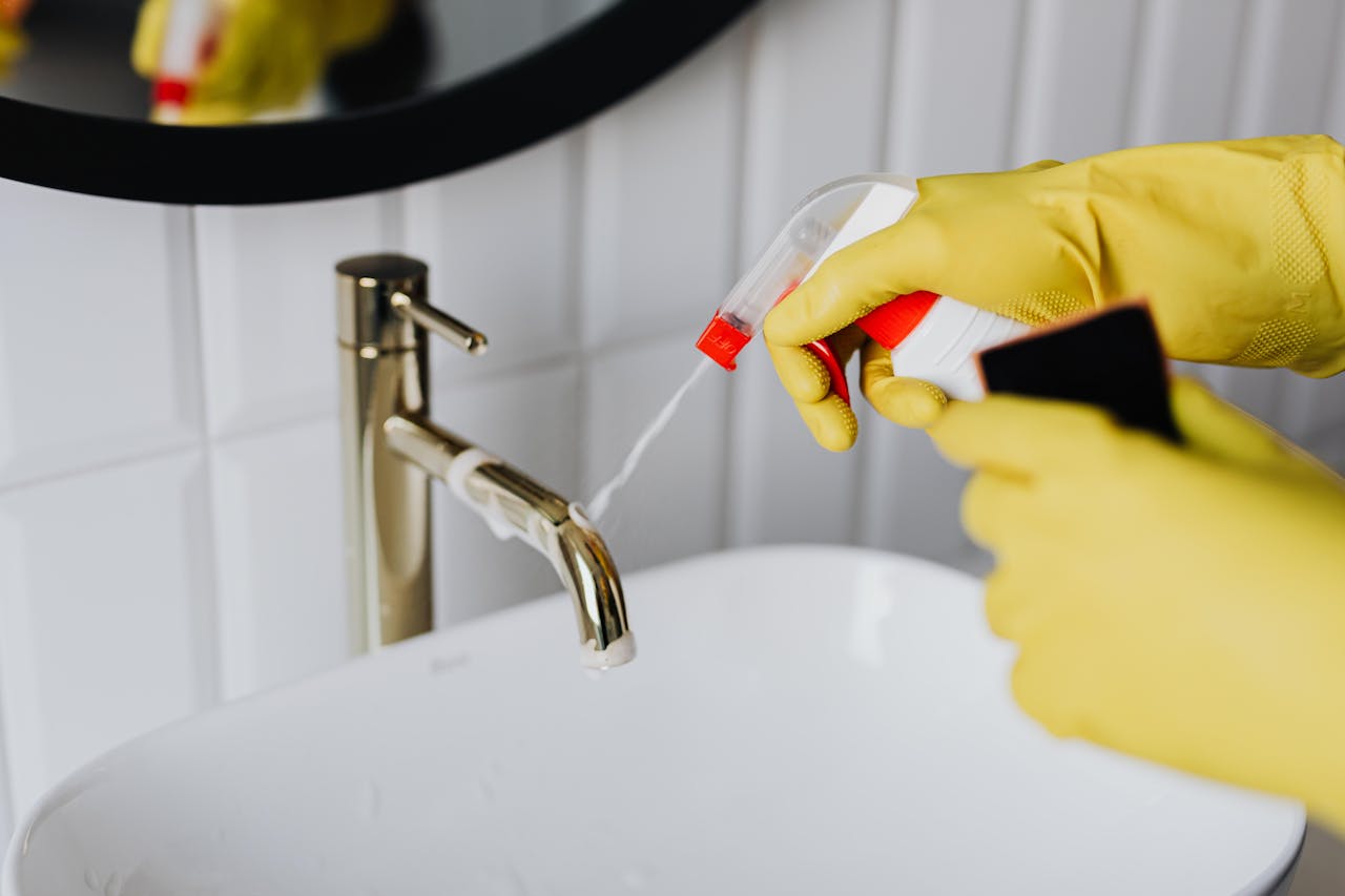 hero-img-02 From above anonymous housekeeper in yellow rubber gloves spraying cleaner to chrome water faucet in bathroom
